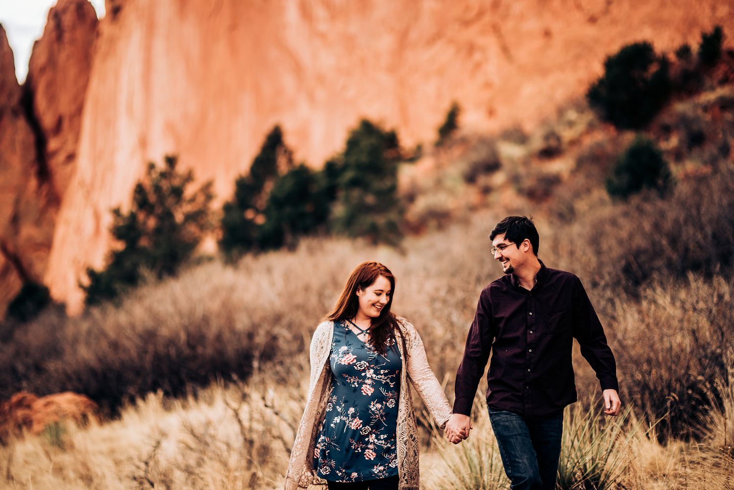 Couple walking hand in hand through Garden of the Gods with red rock formations in background.