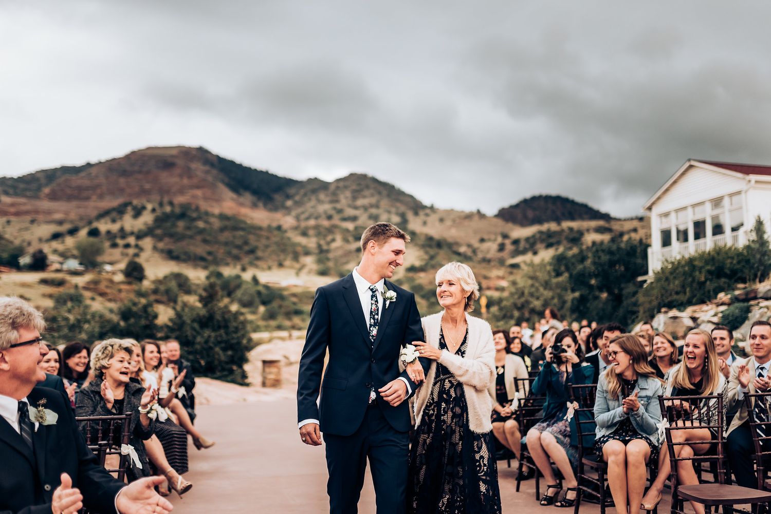 Wedding ceremony guests seated outdoors with mountains and cloudy skies in the background.