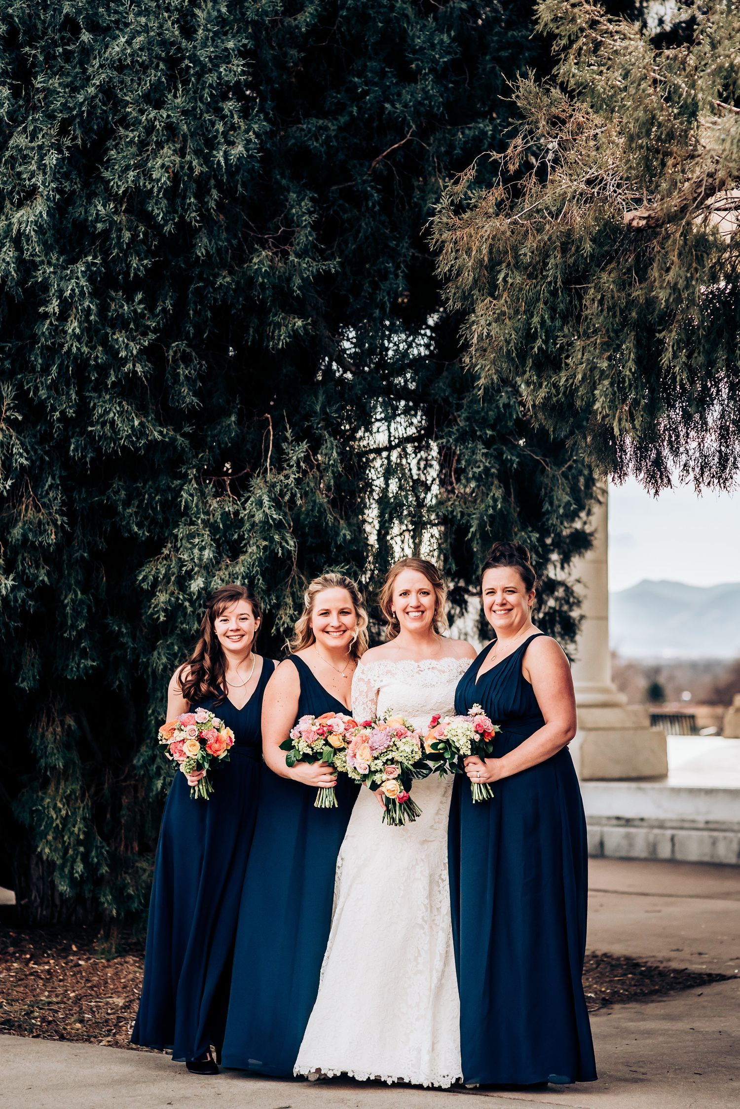 Bridal party with navy blue dresses and pink bouquets pose against evergreen trees.