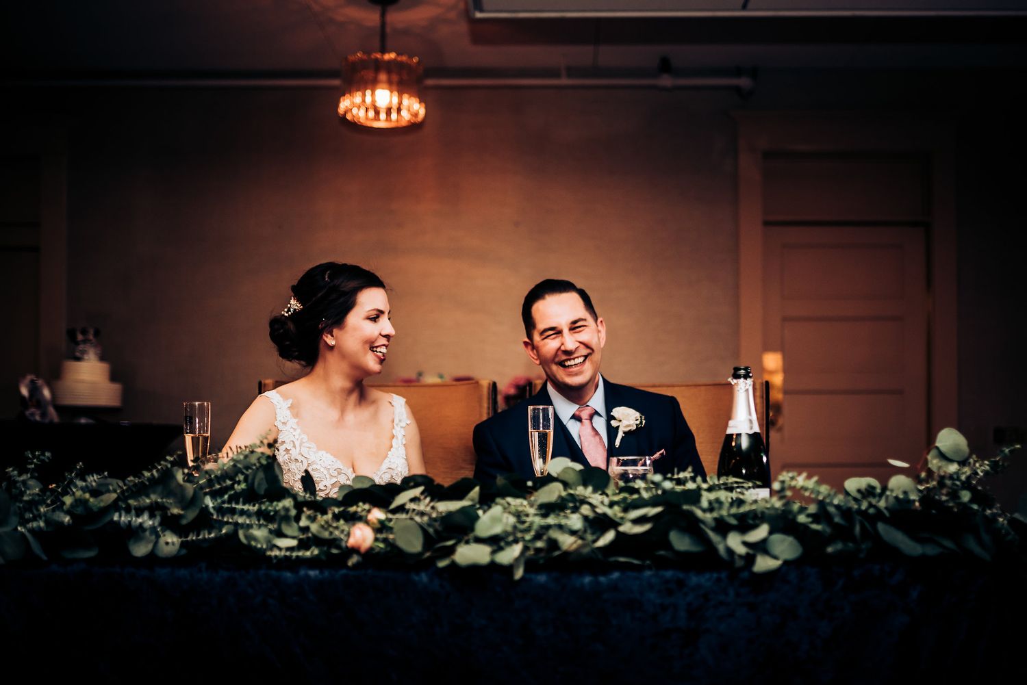 Couple seated at sweetheart table with greenery garland and moody lighting.