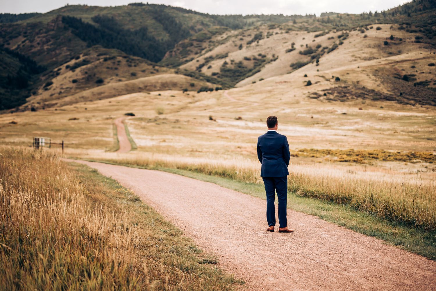 Solo figure standing on dirt path amid rolling golden hills and mountain landscape.