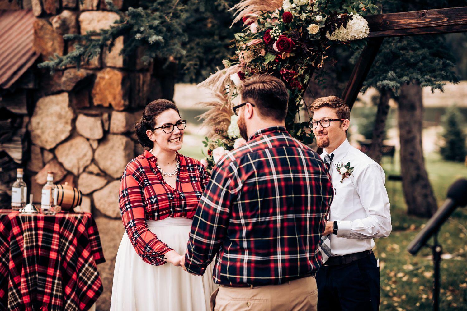 A couple wearing matching plaid shirts exchanges vows during a rustic outdoor wedding ceremony.