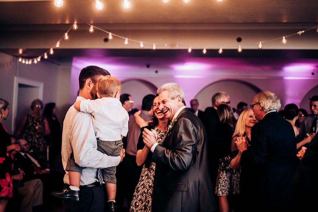 Wedding guests dance and socialize during an evening reception with warm lighting and arched doorways.