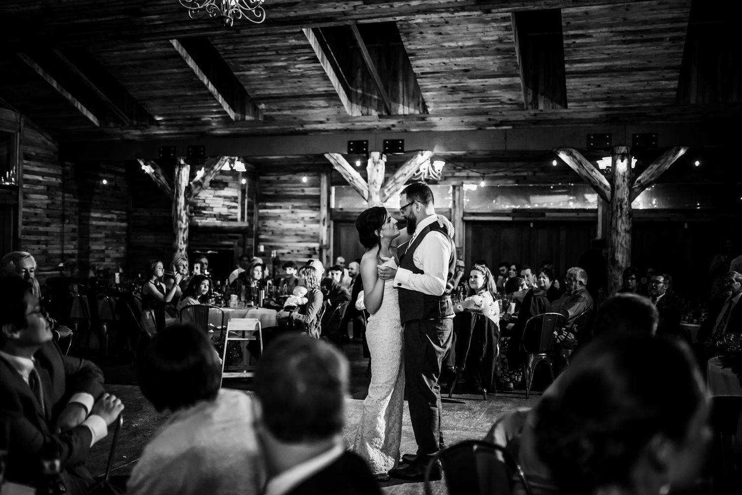 Black and white photo of a couple dancing in a rustic barn surrounded by wedding guests.