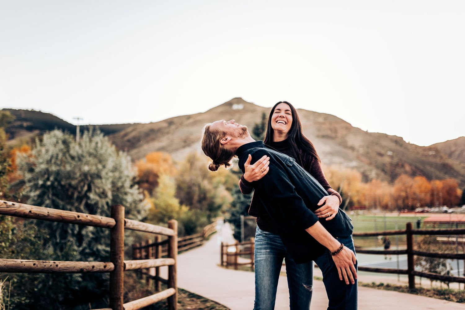 A joyful couple playing and laughing on a mountain path with wooden fencing.