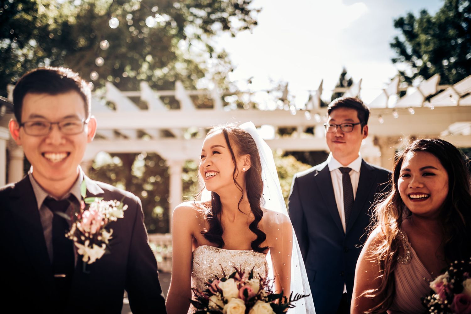 Candid wedding party moment captured outdoors under pergola with natural lighting.