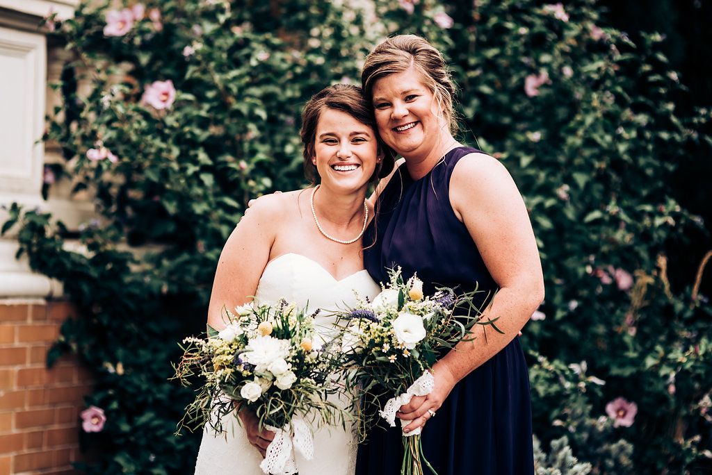 Two people pose together outdoors with white and green wedding bouquets against a floral garden backdrop.