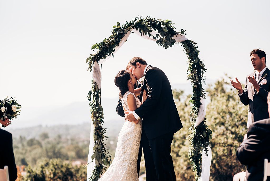 Couple shares first kiss under greenery-adorned wedding arch with mountain views.