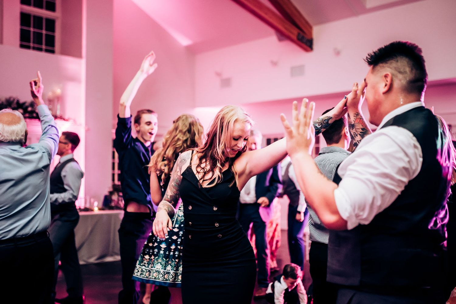 Wedding guests dancing and celebrating with raised hands in a pink-lit reception venue.