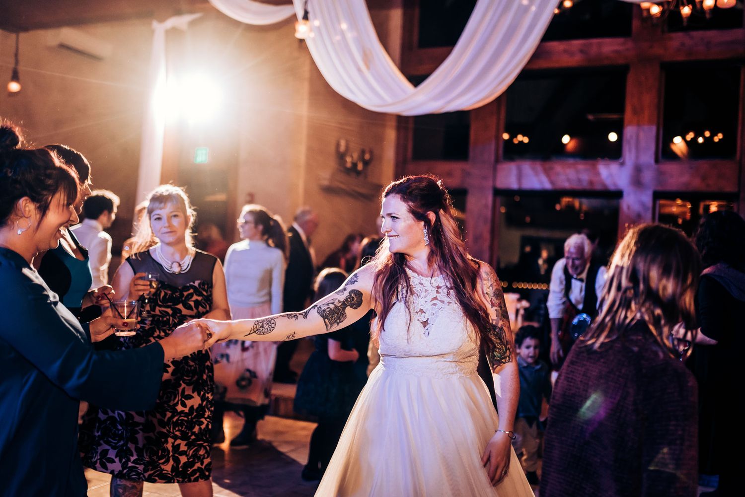 Bride in white dress dancing with guests at a wedding reception under elegant drapes and lighting.