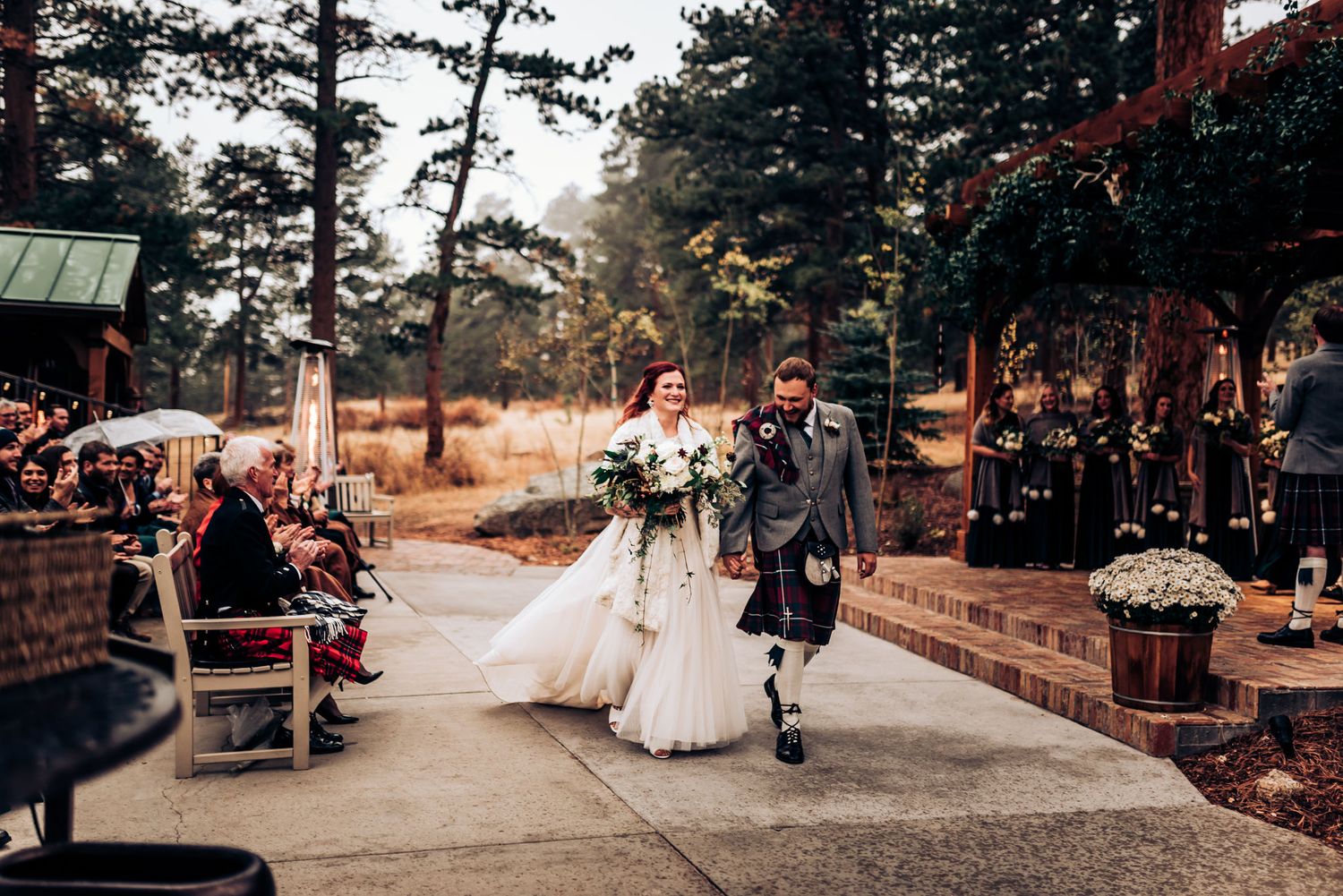 Newlyweds walk down outdoor pathway with Scottish kilt and traditional white wedding dress.