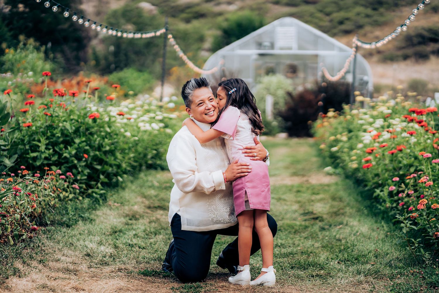 A tender moment in a flower garden path with string lights and a greenhouse in the background.