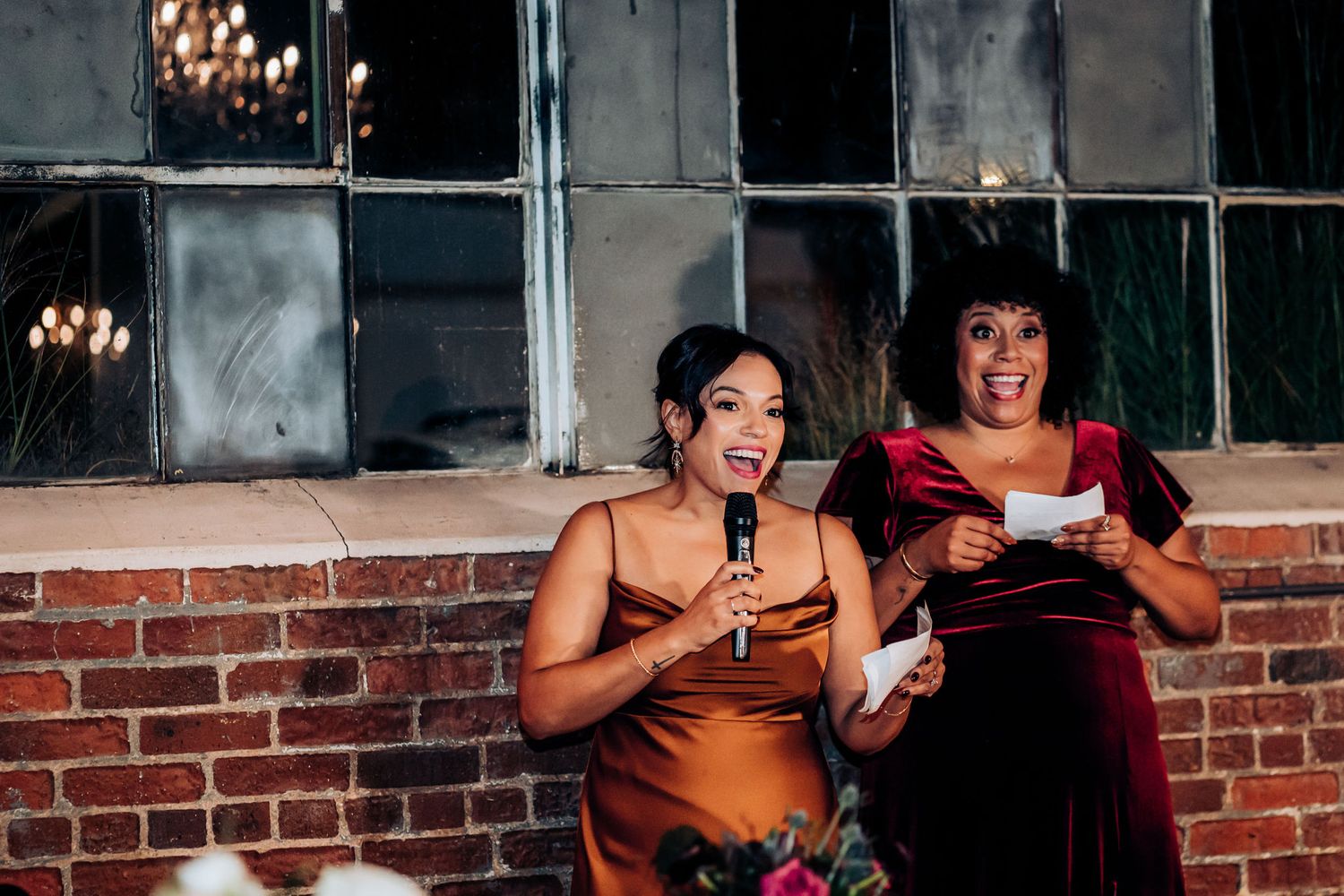 Two speakers share a moment while giving speeches at a wedding reception against a brick backdrop.