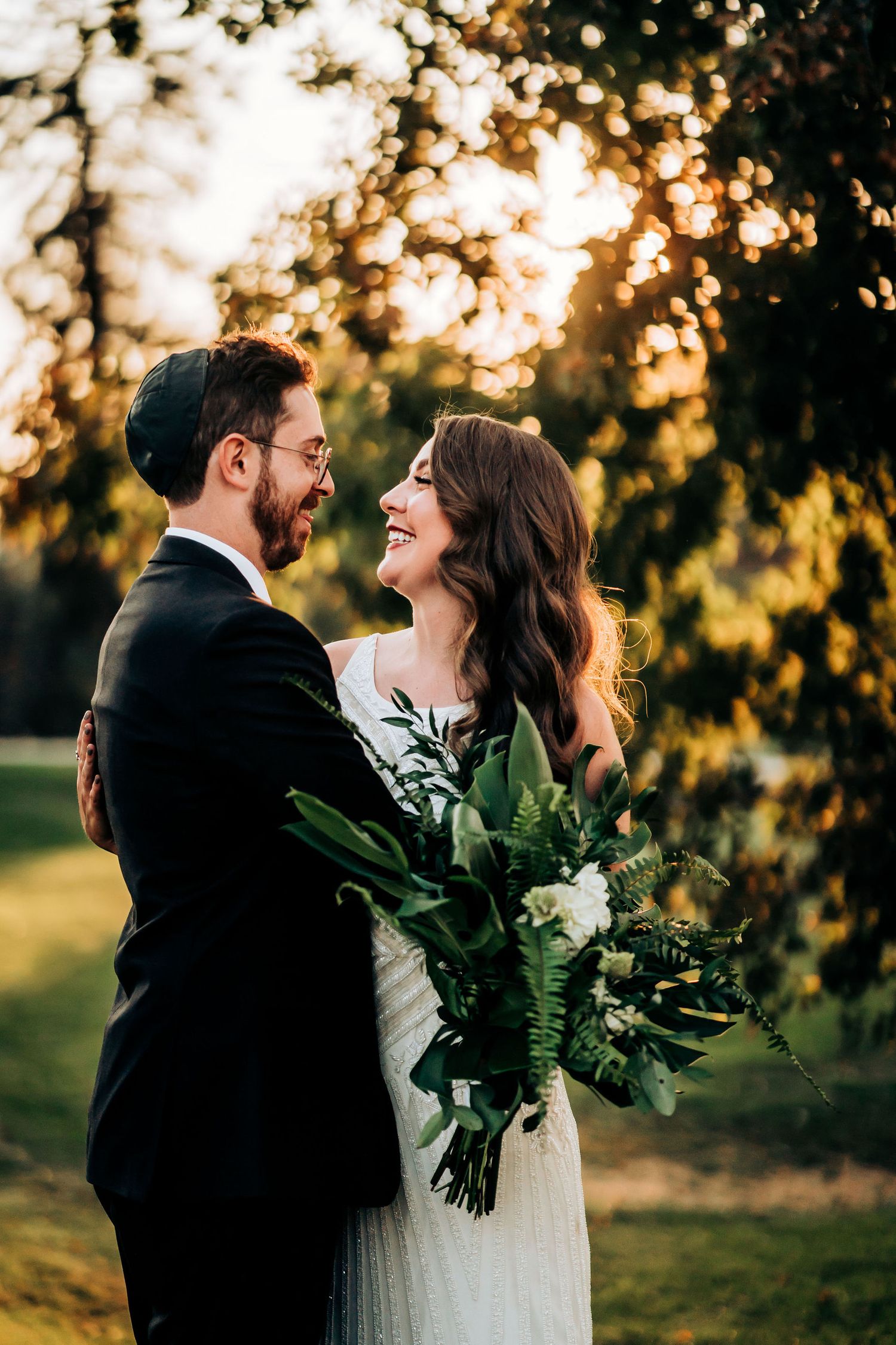 Romantic sunset wedding portrait with couple embracing among trees holding green and white bouquet.
