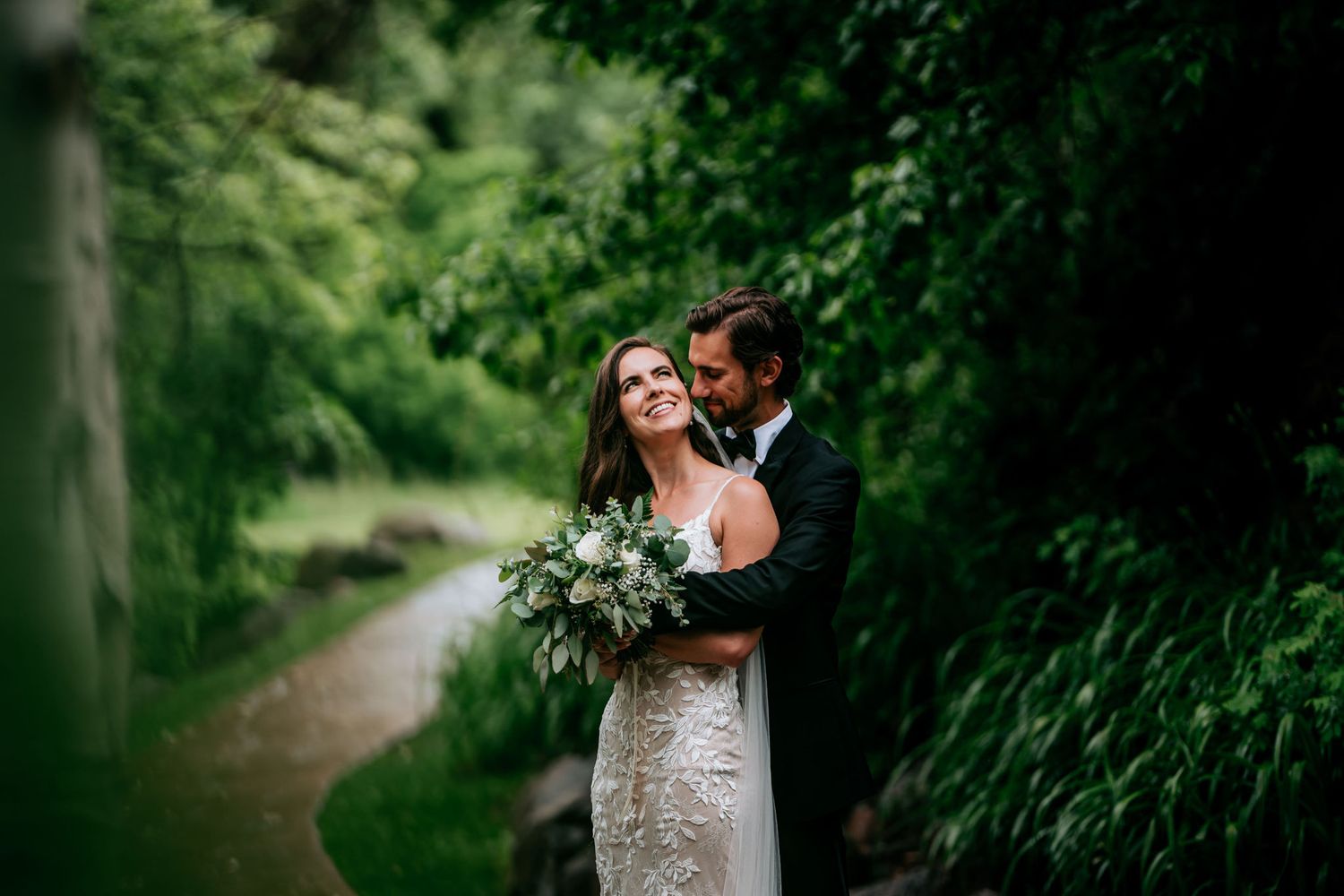 Newlyweds embrace on woodland path with bride holding white and green bouquet.