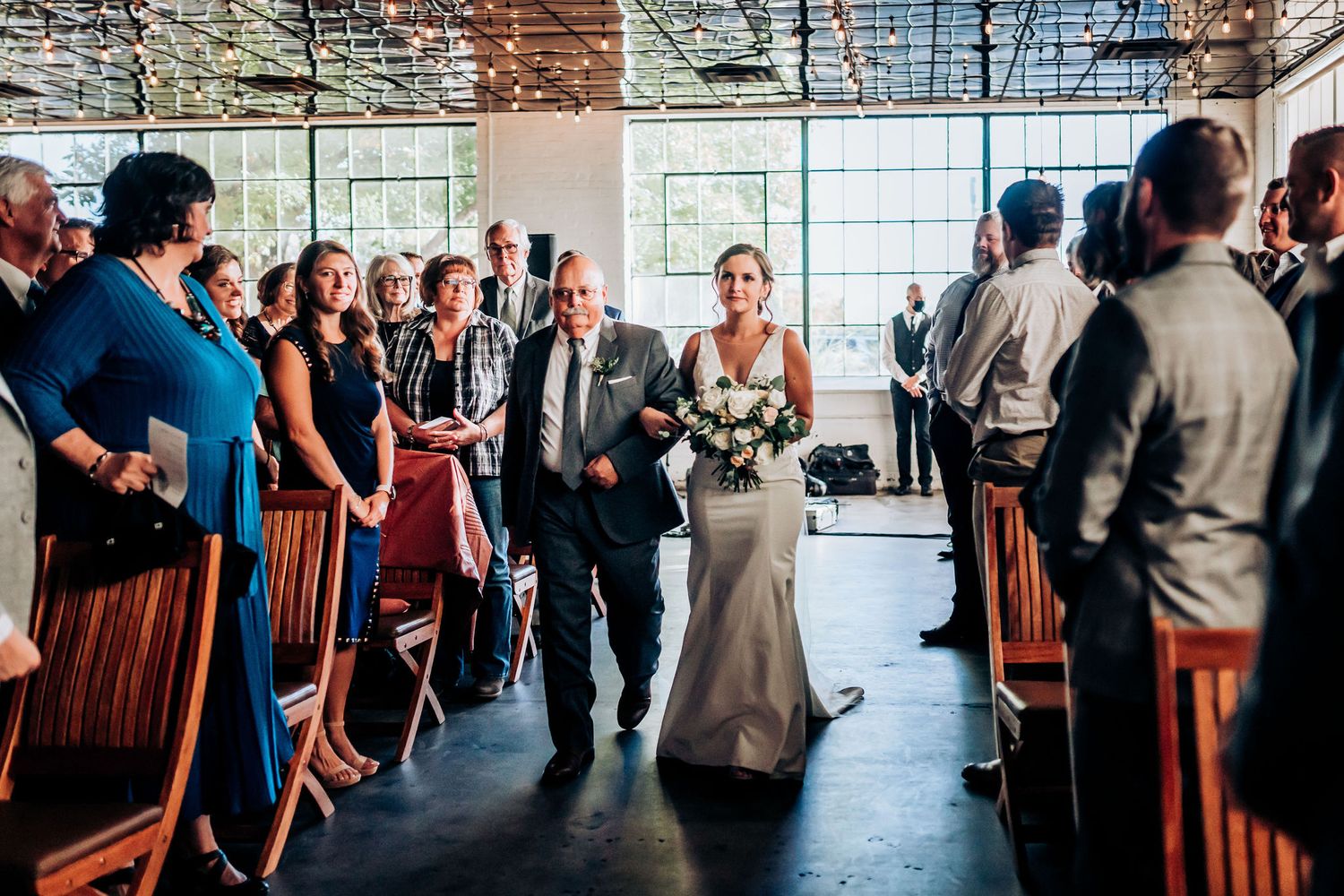 Wedding ceremony processional in an industrial-style venue with guests lining wooden chairs.