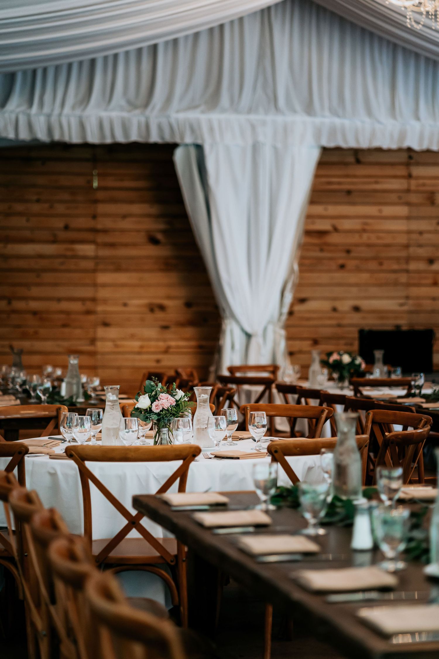 Rustic wedding reception venue with wooden tables, cross-back chairs, and white draping under a tent ceiling.