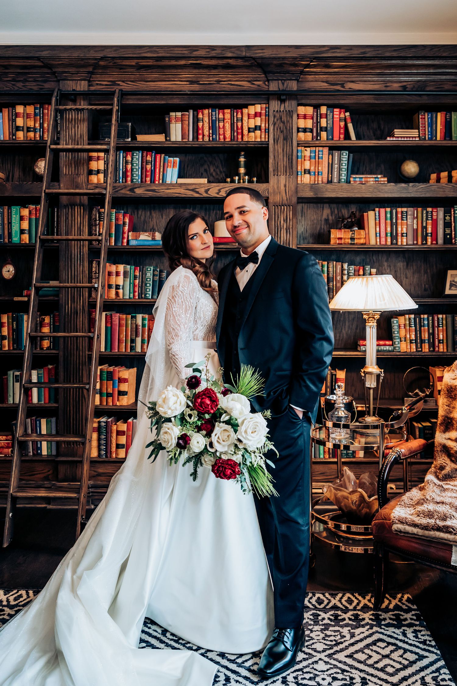 Newlyweds pose in vintage library with floor-to-ceiling bookshelves and elegant bouquet.