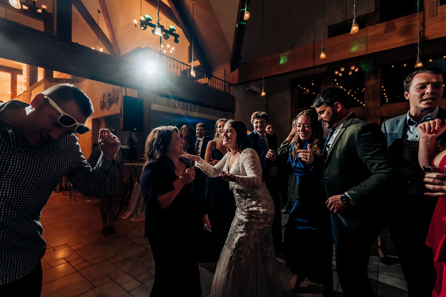 Wedding guests dance and celebrate on a dimly lit dance floor at an indoor reception venue.