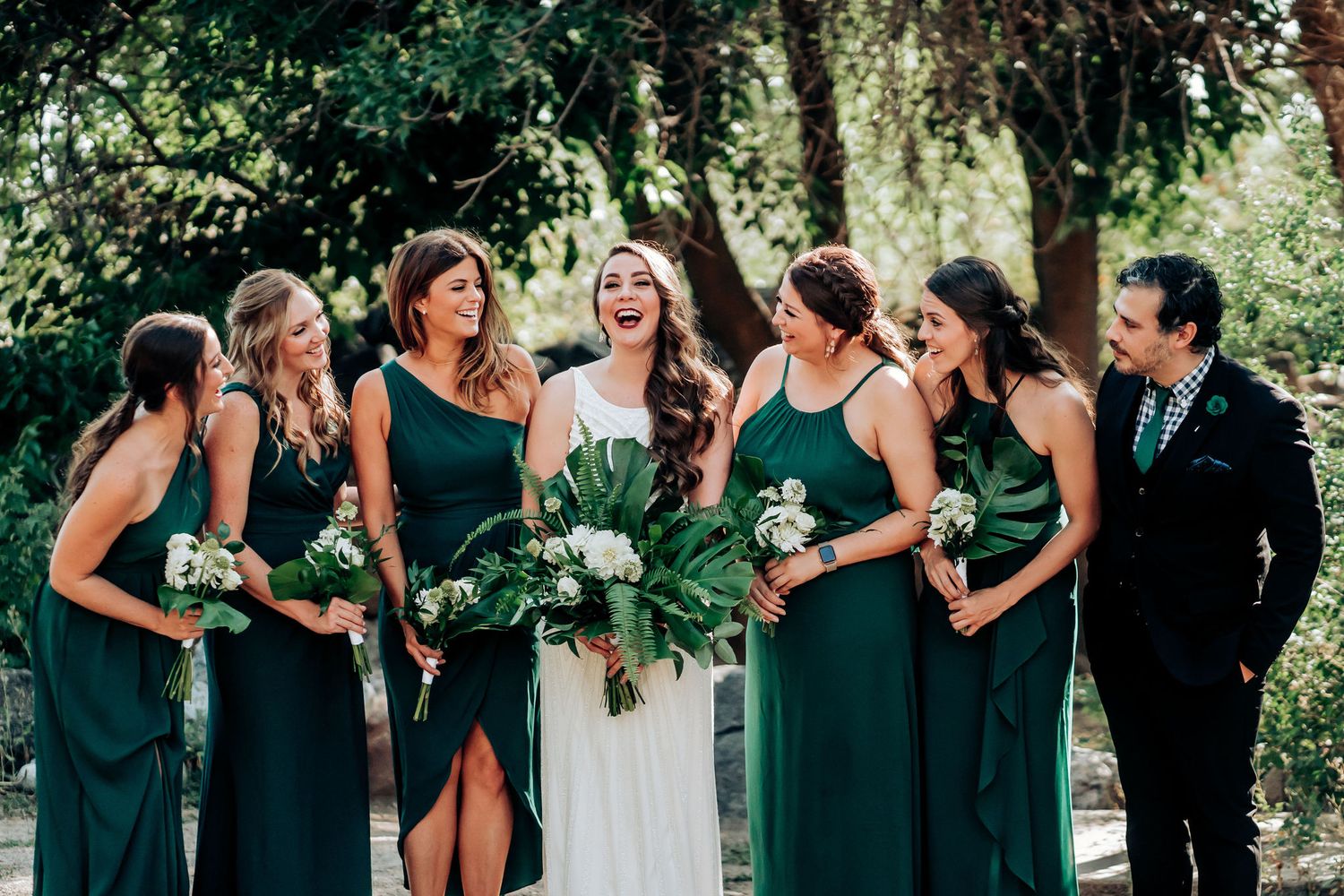 Wedding party shares candid laughs in coordinated emerald green attire outdoors.