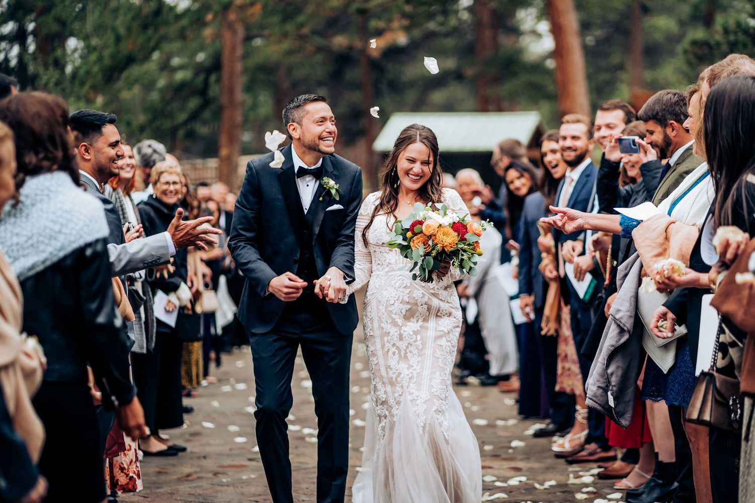 Joyful wedding exit with guests throwing petals along tree-lined pathway.