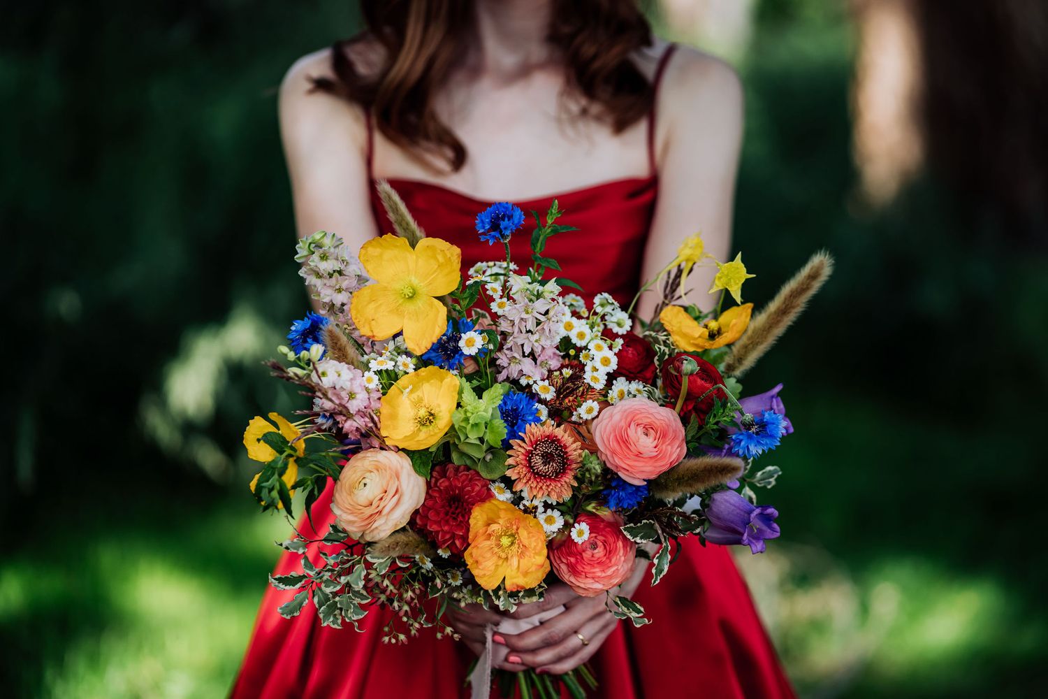 A vibrant wedding bouquet with yellow, pink, and blue flowers held against a red dress.