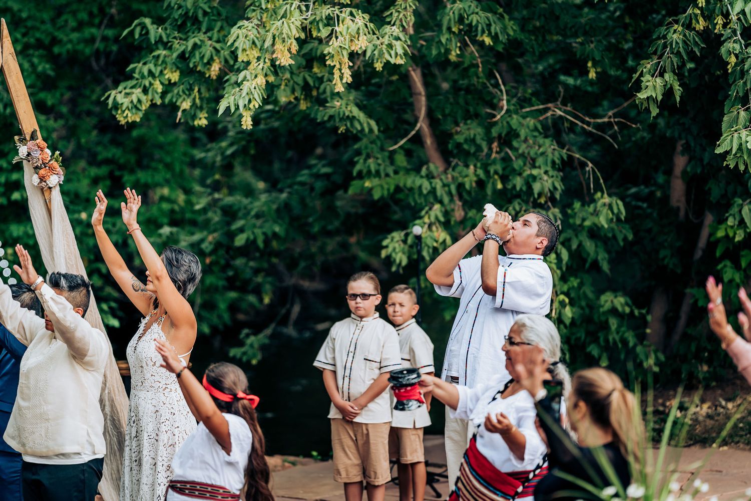 Wedding celebration outdoors with guests raising arms under wooden arch and leafy green trees.