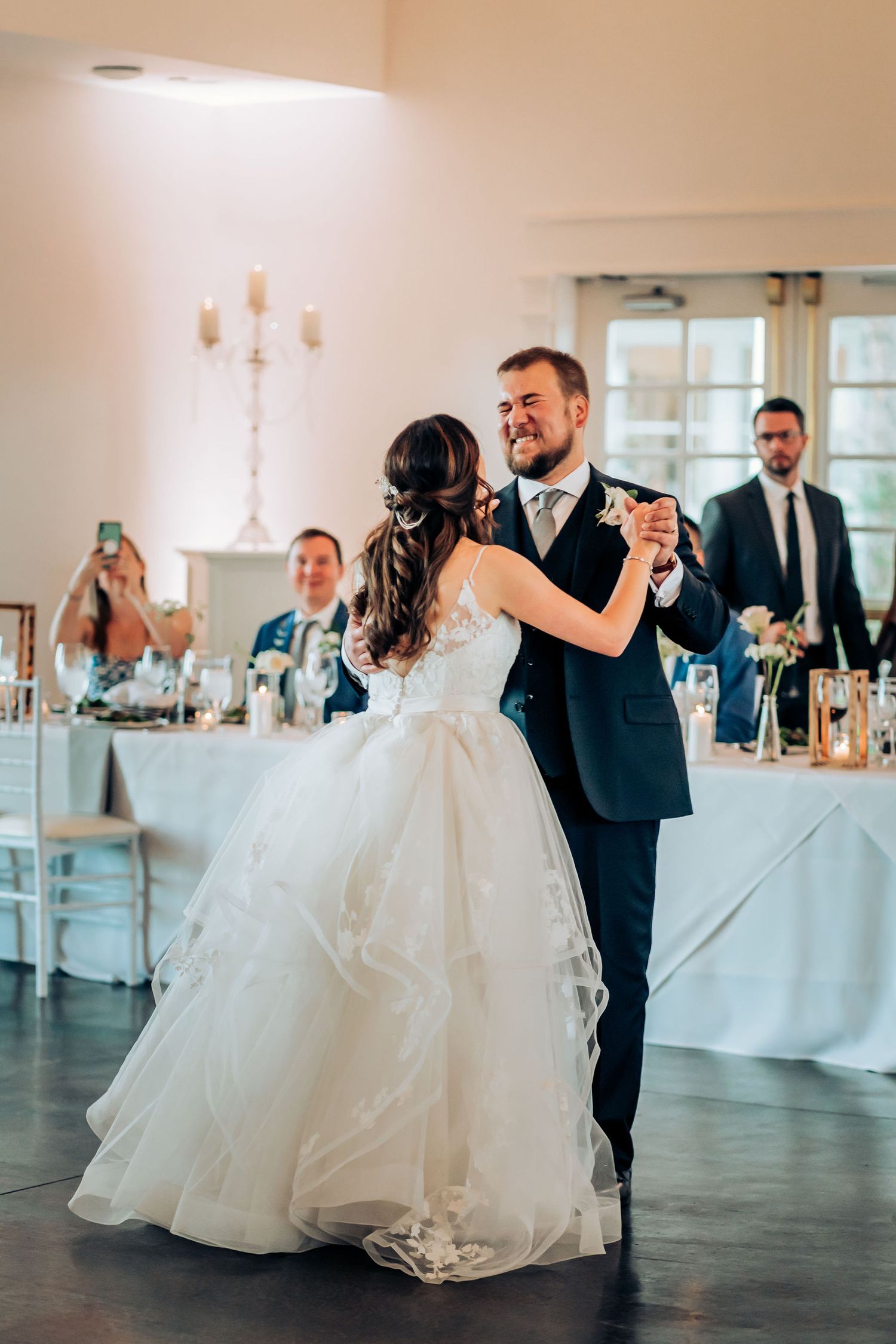 Couple dancing in elegant reception hall with white decor and wedding guests seated at tables.