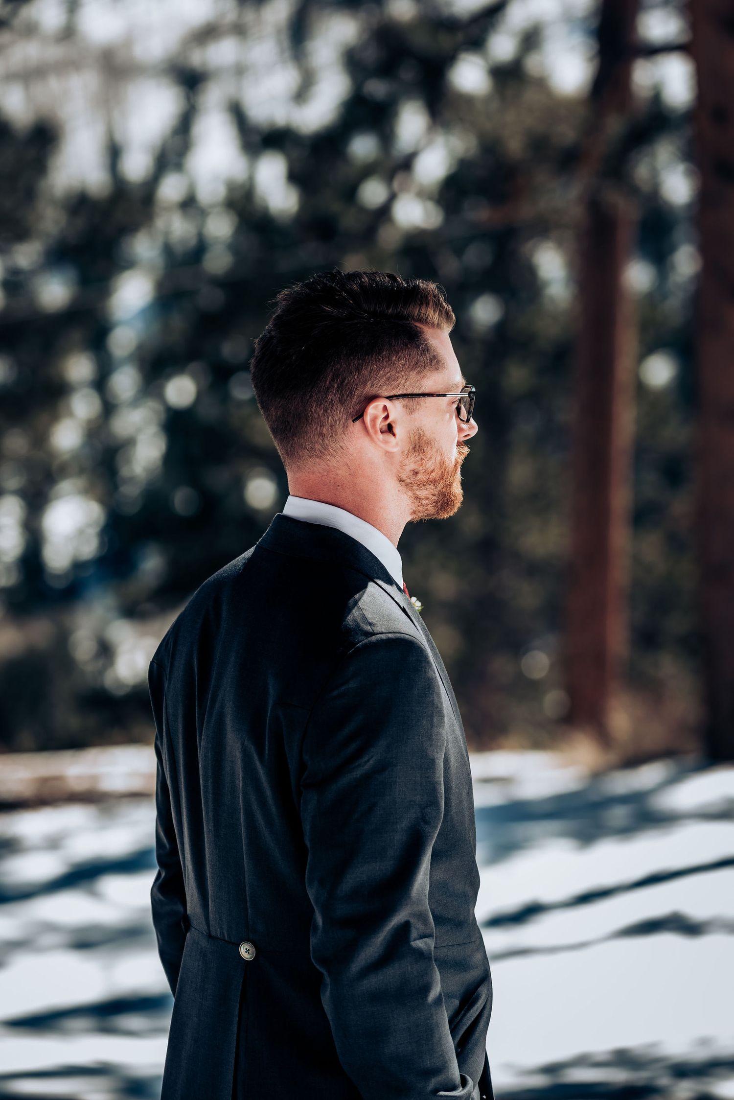 A person wearing a suit and glasses looks to the side in a snowy winter setting.