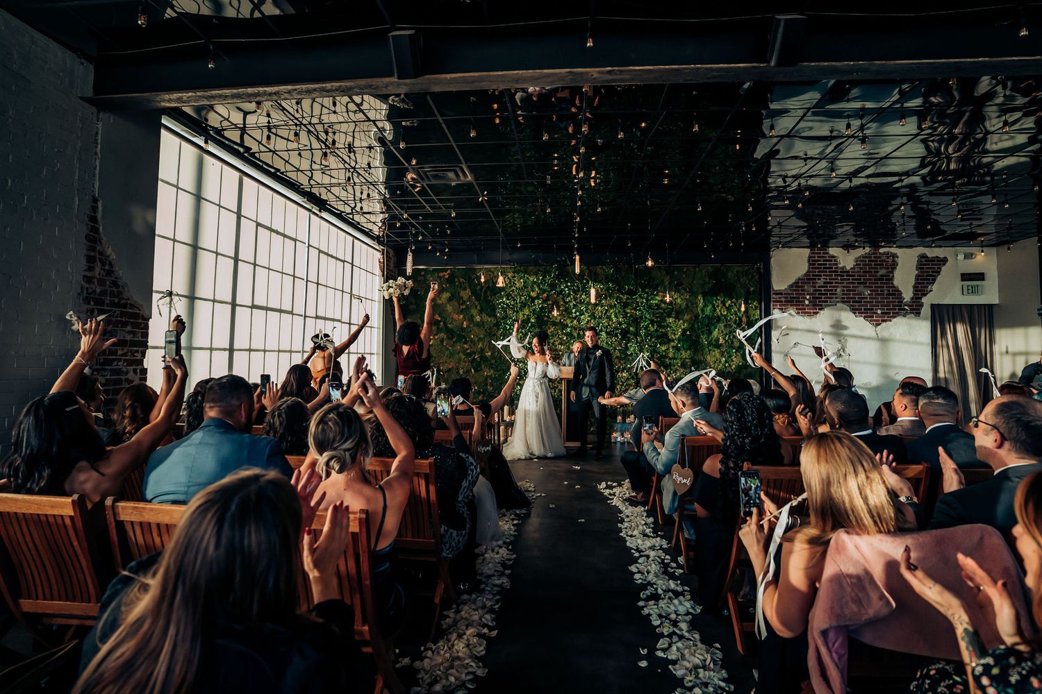 Wedding ceremony guests raise their hands in celebration inside an industrial-style venue with large windows and hanging lights.