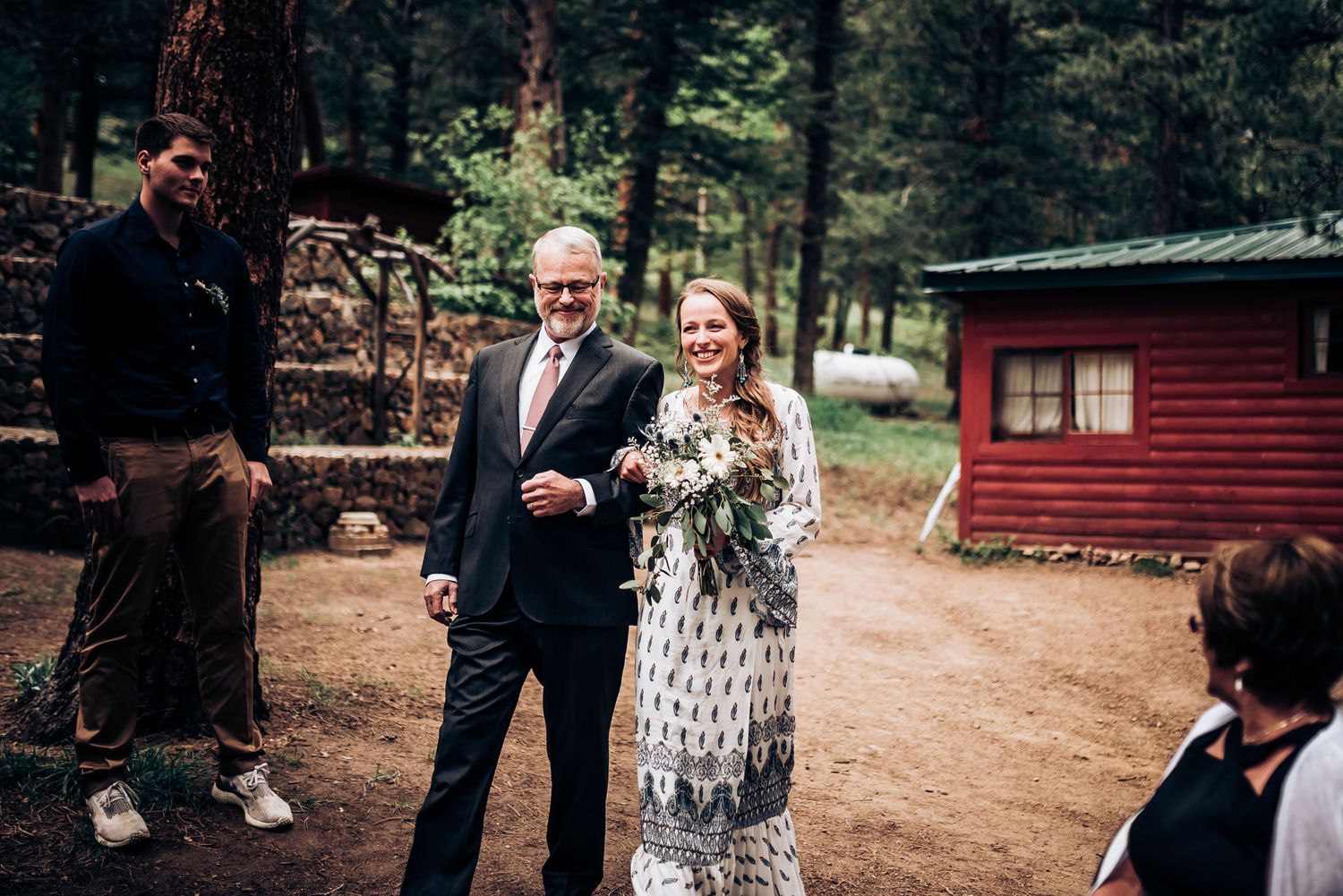 An intimate wedding ceremony takes place on a wooded path near a red cabin.