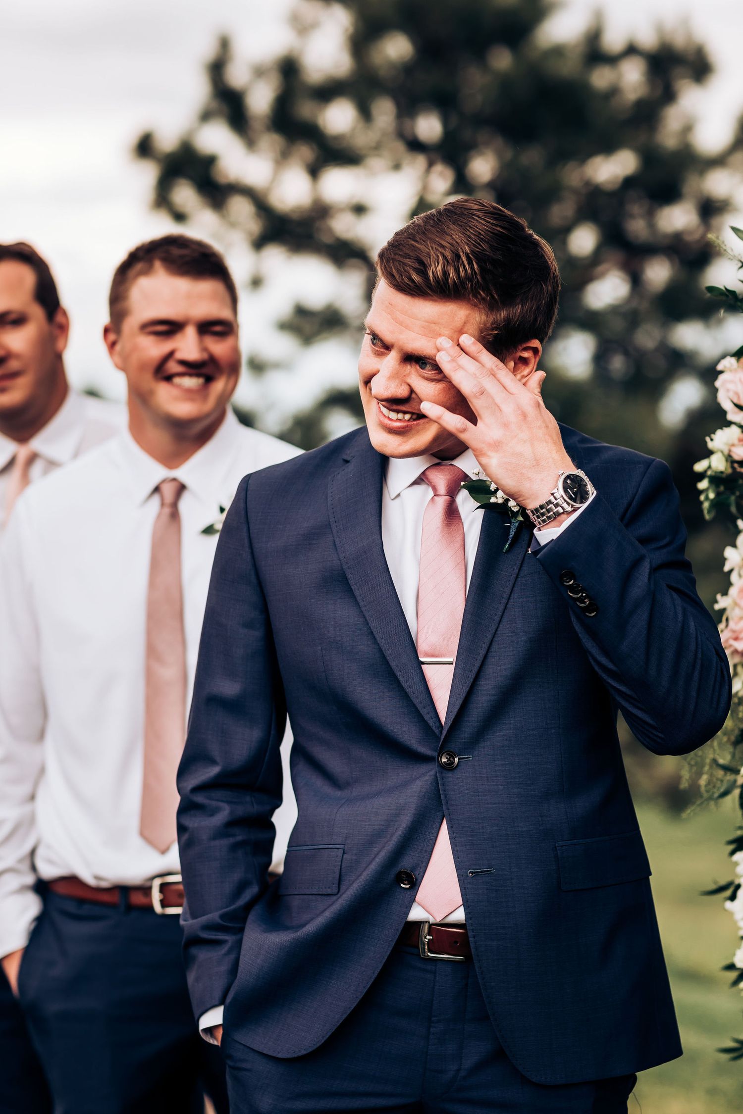 Groomsman in navy suit and pink tie shows emotion during outdoor wedding.
