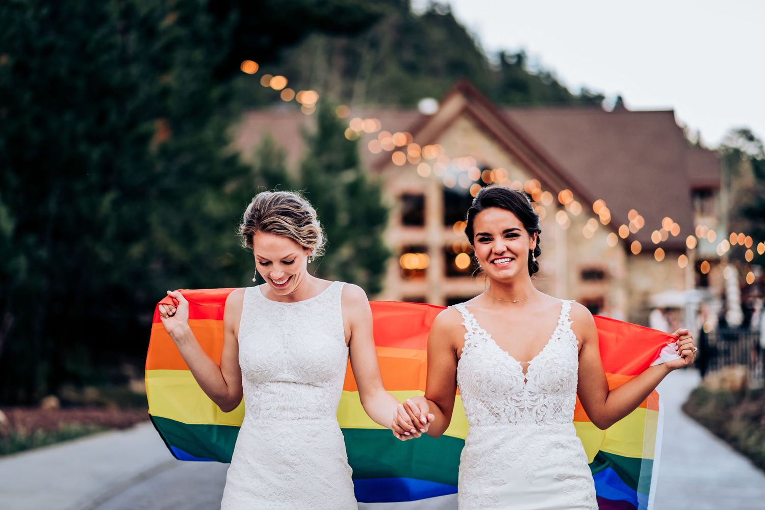 Two people in white wedding dresses hold hands and rainbow flags on a path with lit buildings in background.
