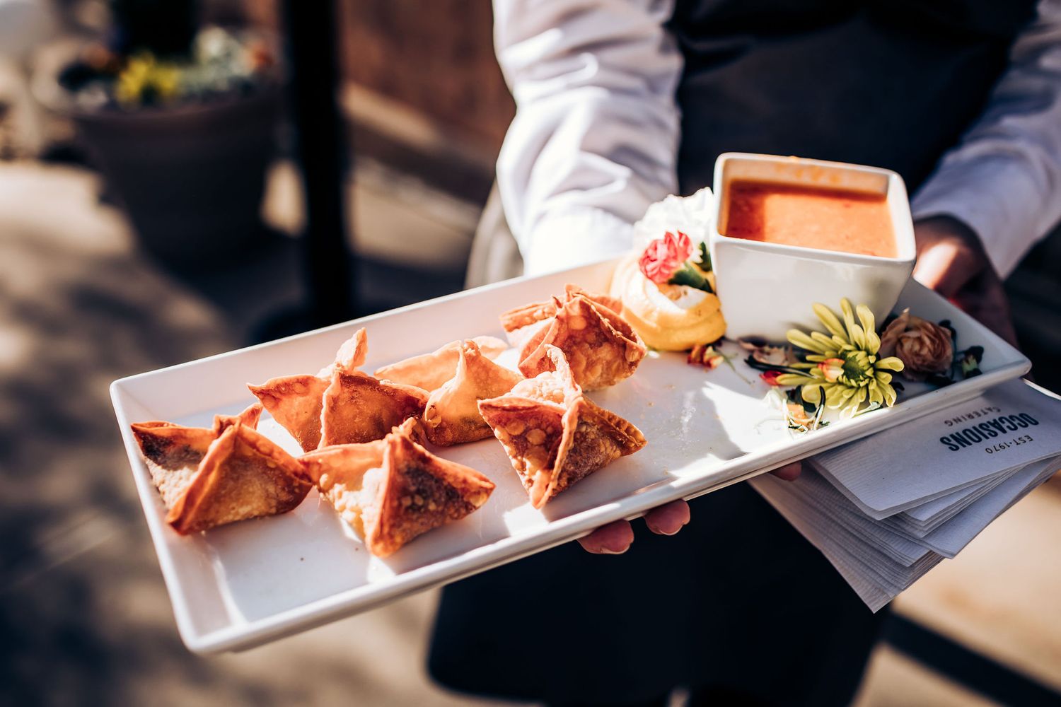 Catering server holds white platter of appetizers including small sandwiches and garnishes.