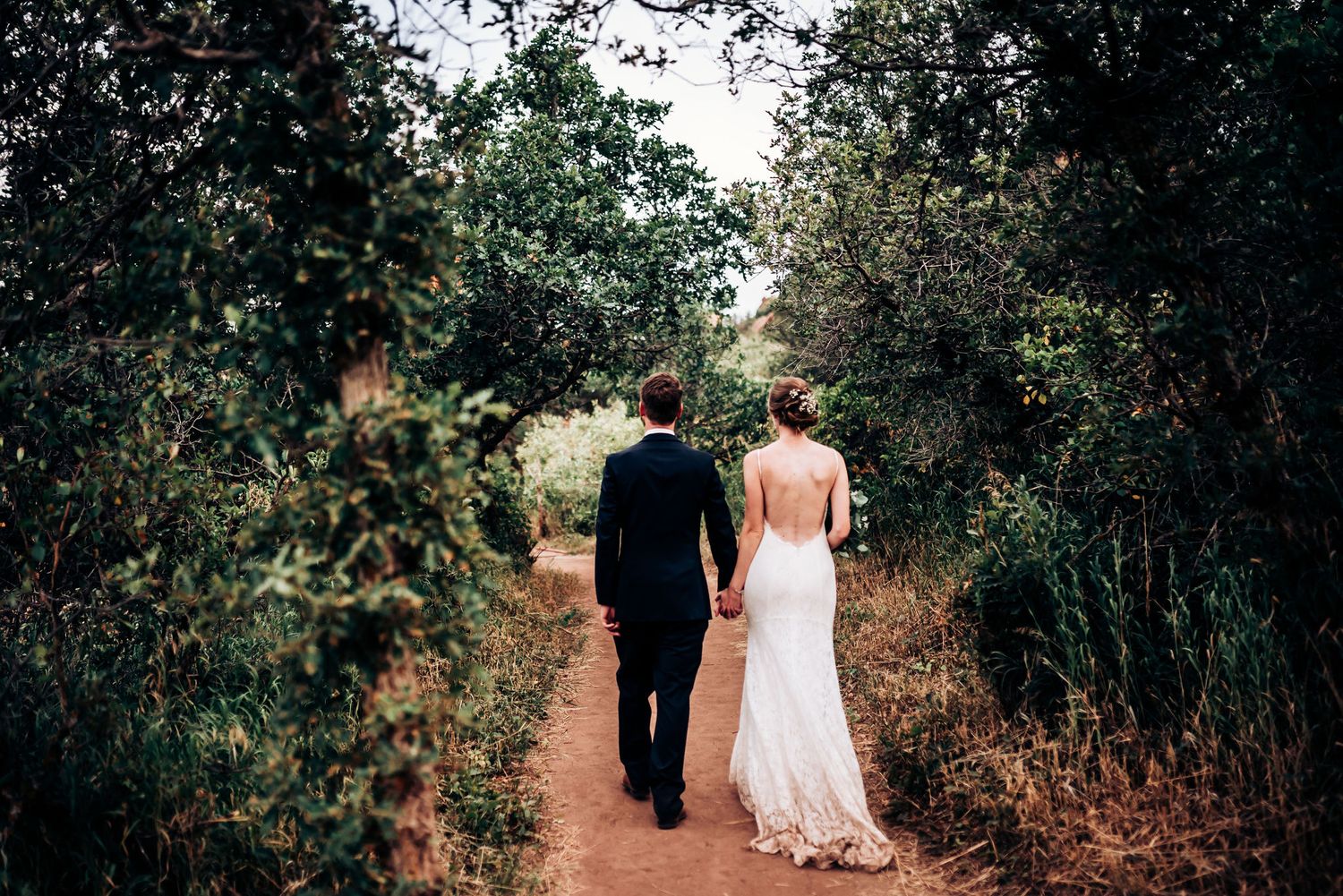 Newlyweds walk hand in hand down a tree-lined dirt path in their wedding attire.