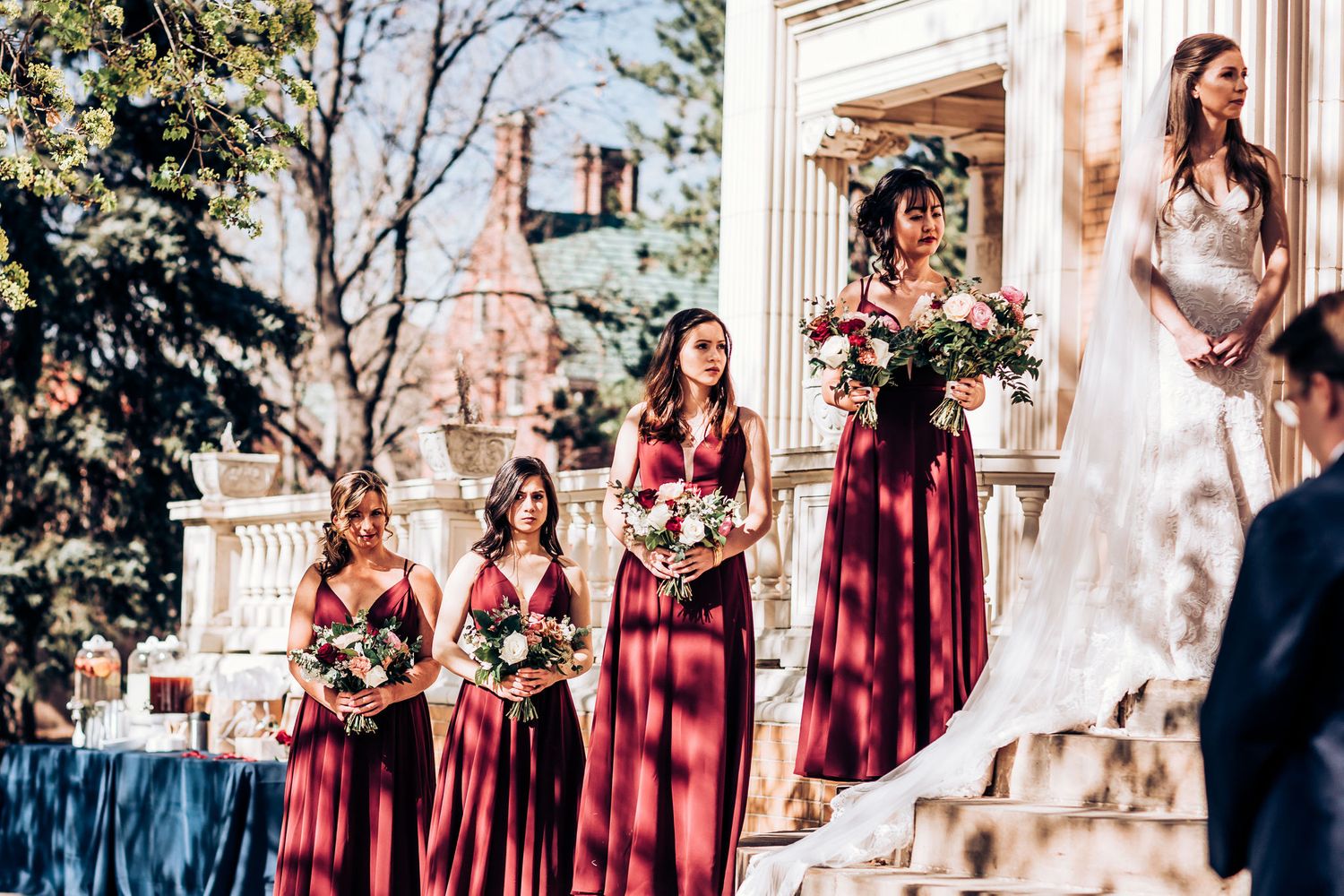 Bridesmaids in burgundy dresses standing on stone steps during an outdoor wedding ceremony.