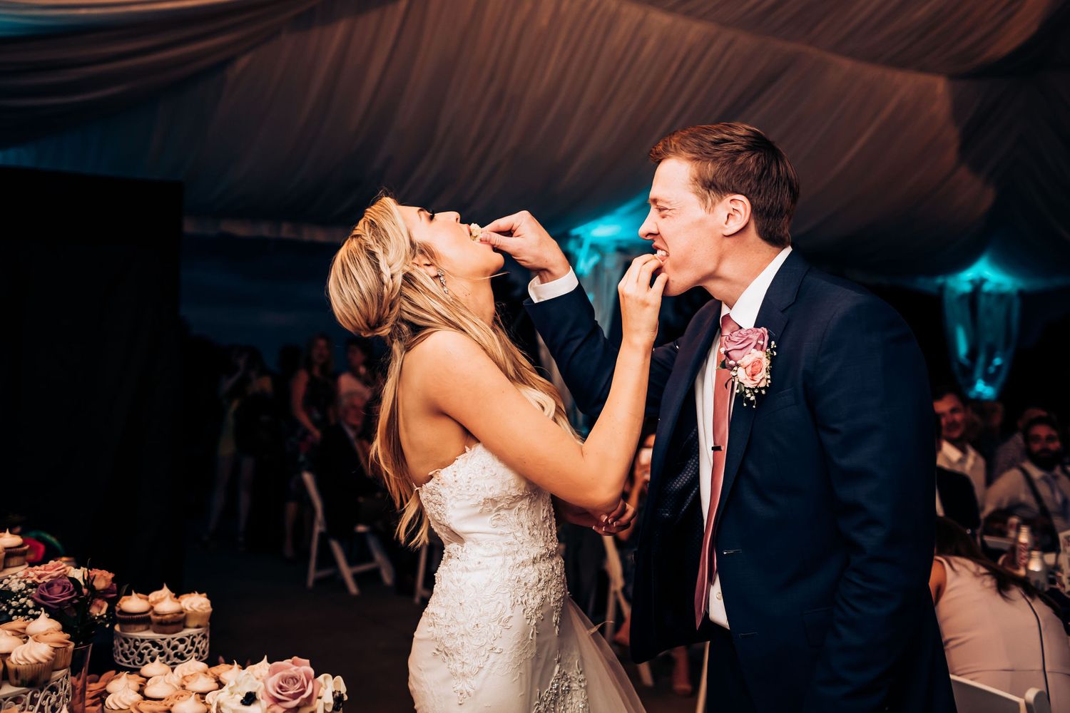 A playful cake-feeding moment during a wedding reception under a draped tent ceiling.