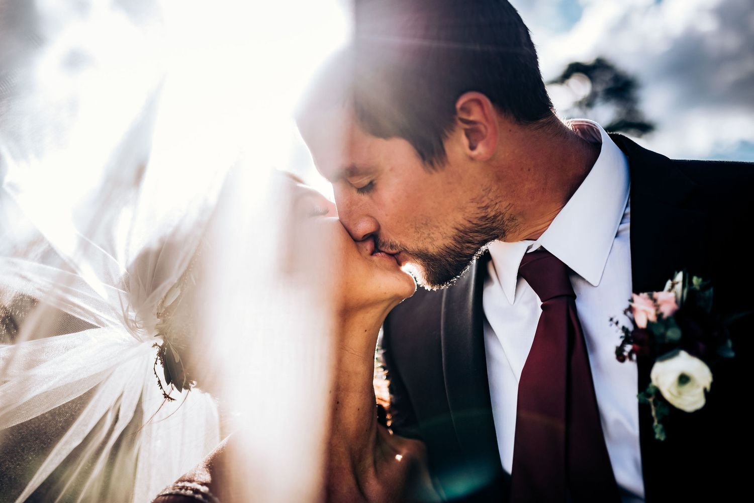 Sun flare illuminates tender wedding moment between couple with flowing veil and burgundy tie.