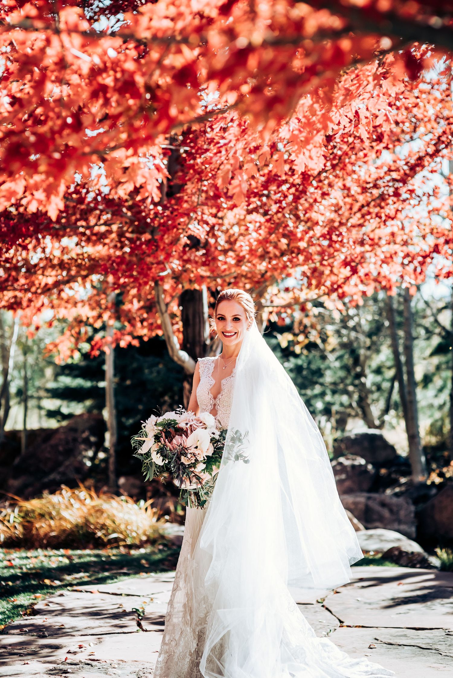 Elegant bride poses with floral bouquet under dramatic red autumn foliage.