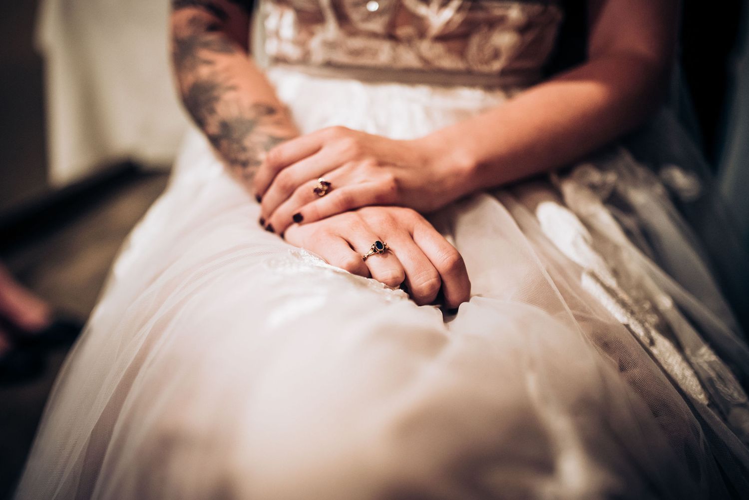 Close-up of tattooed hands resting on white wedding dress fabric.