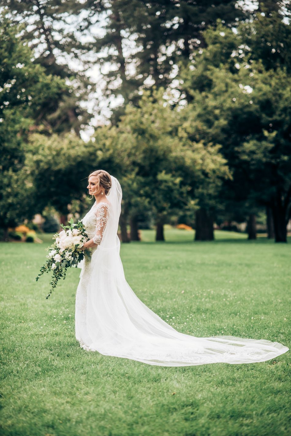 Bride in flowing lace gown and train poses against lush garden landscape.