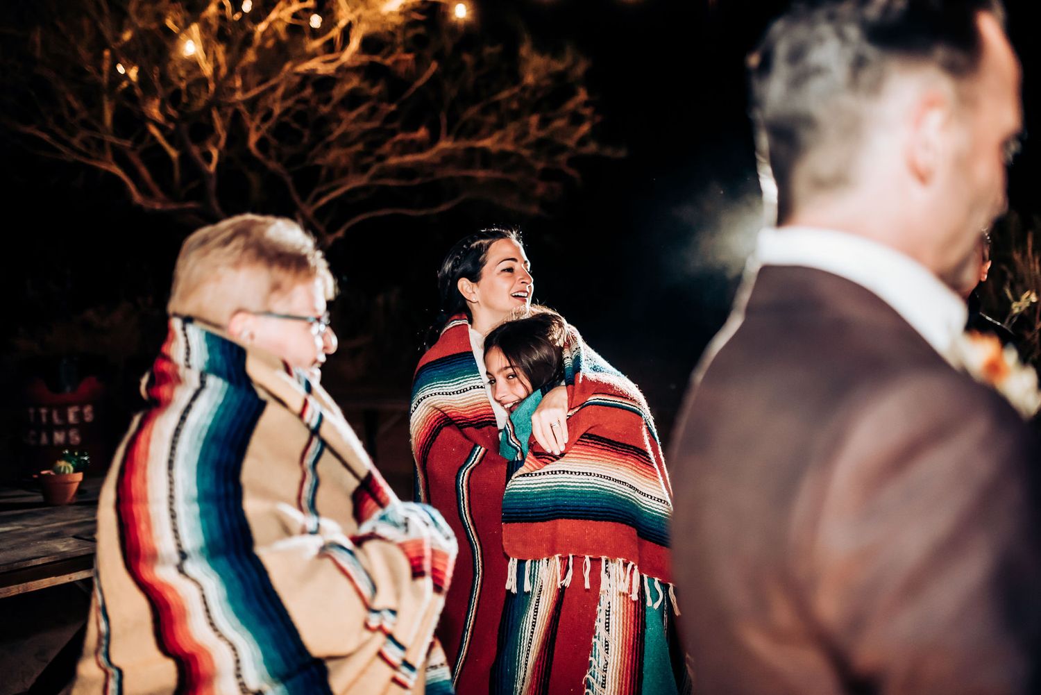 A group wrapped in colorful striped blankets gather outdoors at night under string lights.