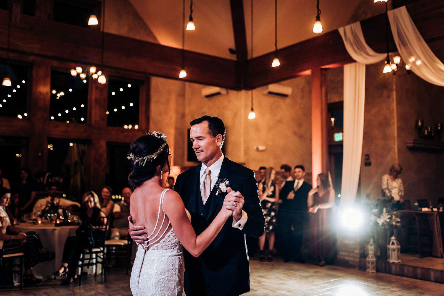 A couple dances together during their wedding reception in a warmly lit rustic venue.