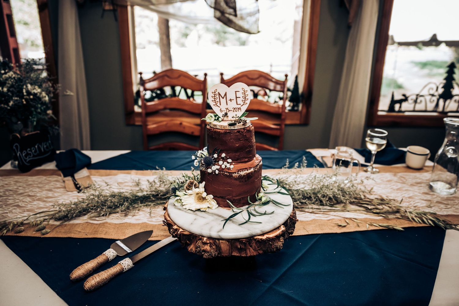 Rustic wedding cake display with greenery on wooden stand against navy tablecloth.