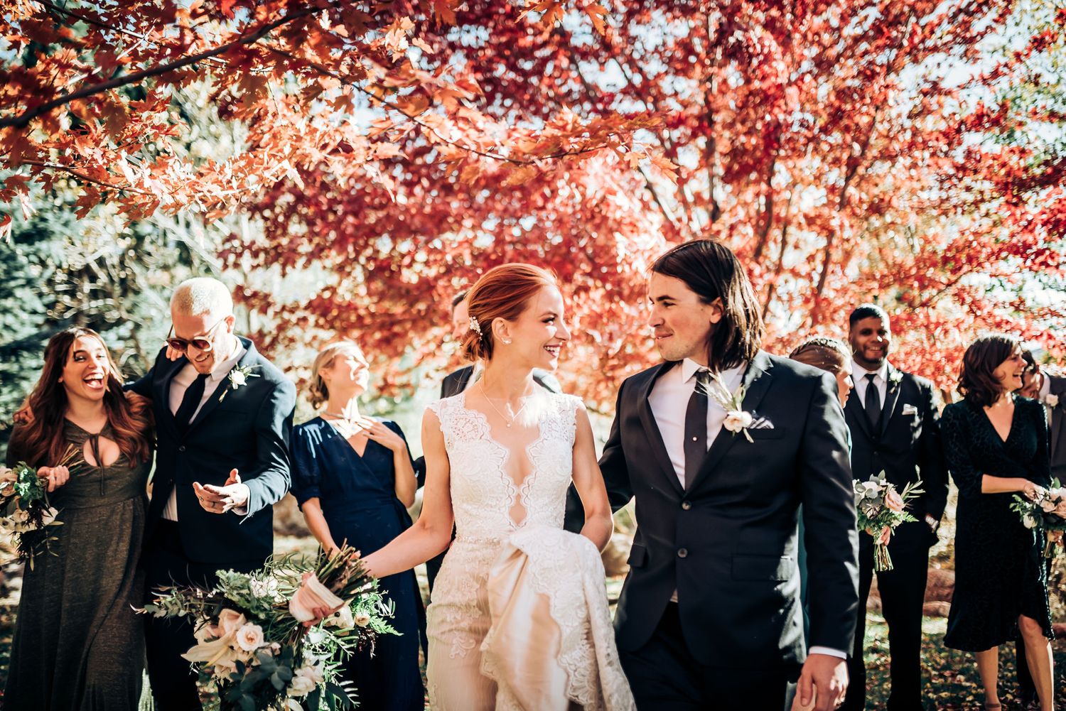 Wedding party walking under vibrant red autumn trees during outdoor celebration.