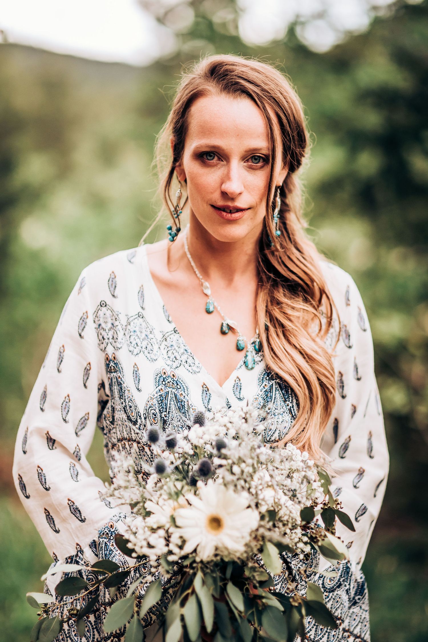 Bride in white patterned dress holds bouquet while posing in outdoor setting.