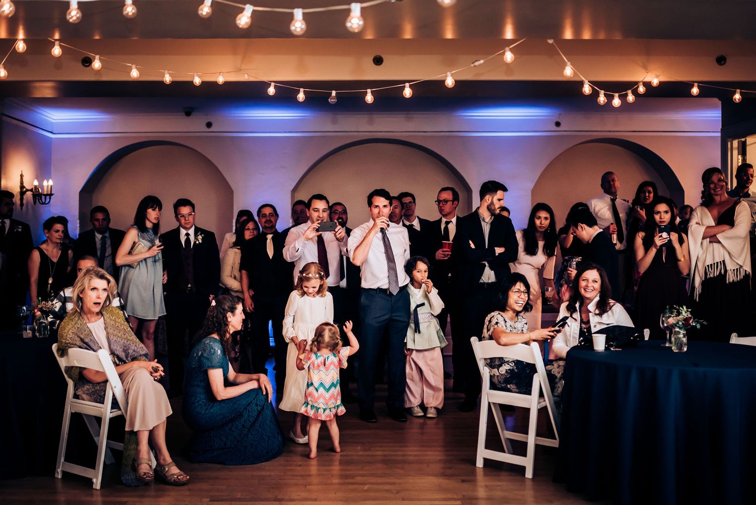 Wedding celebration in an elegant ballroom with string lights and arched windows.