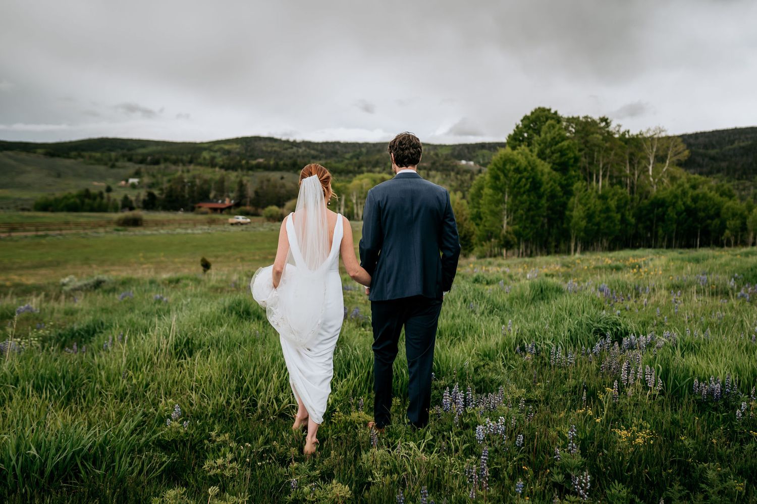 Couple walking through mountain meadow with flowing white dress and navy suit.