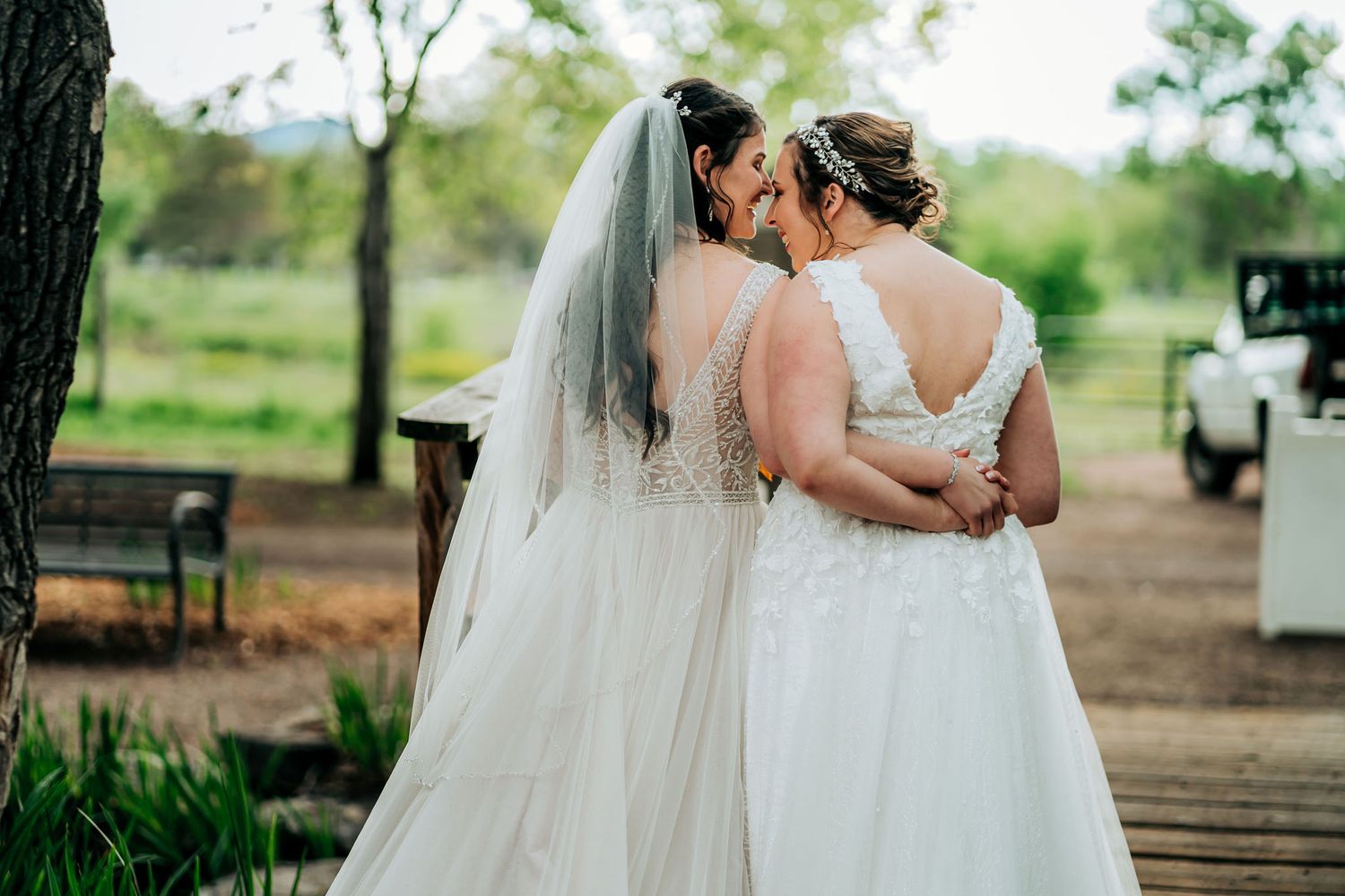 Two people in white wedding dresses embrace outdoors under trees on a rustic path.