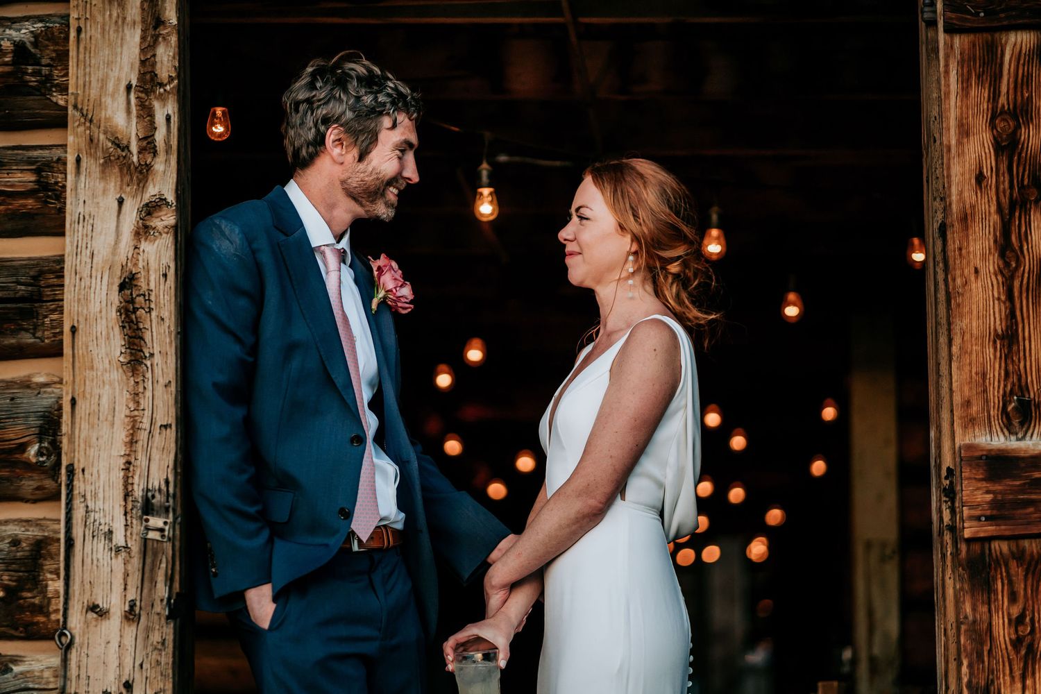 A couple shares an intimate moment in a rustic barn setting with glowing string lights.